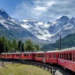 Röda tåg slingrar sig genom en grönskande dal med snöklädda berg i bakgrunden. En drömresa med nattåg genom Europa.