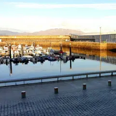 Barcos pesqueros amarrados en el puerto de Finisterre, con un edificio moderno al fondo y montañas en la distancia.
