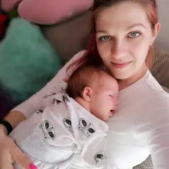 Mama tuli otula swoje śpiące dziecko, czułe kangurowanie dziecka w białym pajacyku z motywem zwierzątek.