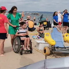 Na plaży dla niepełnosprawnych ratowniczka pomaga dziewczynce na wózku inwalidzkim. W tle widać innych plażowiczów i morze.
