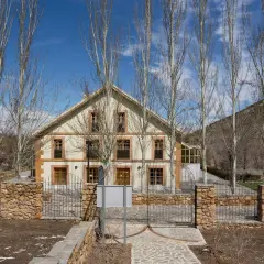 Edificio de piedra y madera con árboles secos delante. El camino de entrada está pavimentado con piedras. Las menas de la montaña se ven al fondo.