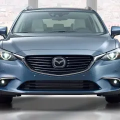 Niebieska Mazda 6 GJ kombi z przodu, z włączonymi światłami, na tle miasta.