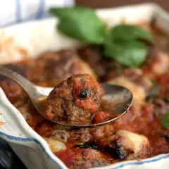 En sked lyfter en köttbulle täckt av mustig tomatsås ur en ugnsform. En härlig pasta med tomatsås väntar!
