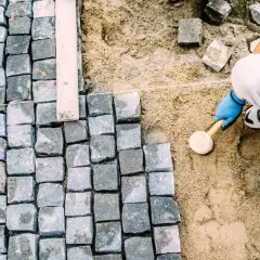 Pracownik w niebieskich rękawiczkach ubija kostkę brukową gumowym młotkiem, tworząc równą nawierzchnię.