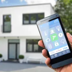 Dłoń trzyma smartfon z aplikacją do **instalacji inteligentnego domu**. Na ekranie widoczne ikony sterowania oświetleniem, ogrzewaniem, kamerami i innymi funkcjami.