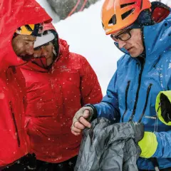 Bergsteiger im Schnee, die sich über Ausrüstung beugen. Die Kleidung, darunter eine rote Jacke mit "Arc'teryx" Schriftzug, zeugt von hoher Qualität und Herkunft für extreme Bedingungen.