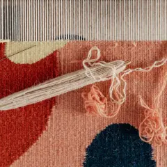 Wzorzysty gobelin w trakcie tworzenia. Widać fragmenty w kolorach czerwonym, łososiowym i granatowym. Użyto naturalnego materiału ramia.