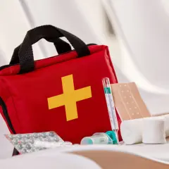 Czerwona apteczka turystyczna w góry, z żółtym krzyżem. Obok termometr, plastry, bandaże i leki.