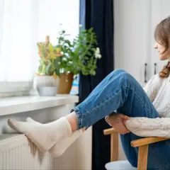 Kobieta w swetrze i jeansach siedzi przy oknie, grzejąc stopy w skarpetkach na kaloryferze. To sposób na obniżanie temperatury w domu.