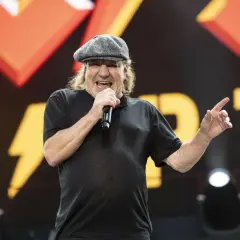 Brian Johnson young, w czapce i czarnej koszulce, śpiewa do mikrofonu na tle sceny z logo zespołu.
