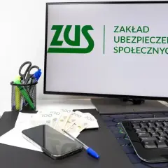 Na biurku leżą pieniądze, telefon i długopis. Na monitorze widnieje logo ZUS, symbolizujące składkę ZUS.