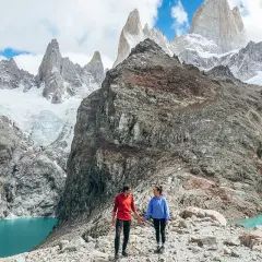 Deux randonneurs se tiennent la main sur un sentier rocheux, admirant un lac turquoise et des montagnes enneigées lors d'une randonnée argentine.