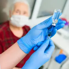 Pielęgniarka w niebieskich rękawiczkach przygotowuje zastrzyk z lekiem, być może z morfiną, dla starszej pacjentki w masce.