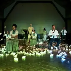 Scena teatralna pełna papierowych ptaków, ludzie w strojach z epoki i współczesnych ubraniach, posąg. To wszystko tworzy atmosferę teatru absurdu.