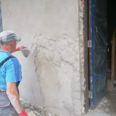 Pracownik tynkuje stare ściany zewnętrzne, nakładając zaprawę kielnią. Widać fragment drzwi i schody w tle.