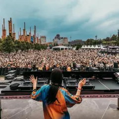 DJ set al futur festival, folla in delirio sotto un cielo nuvoloso, con un'architettura industriale sullo sfondo.