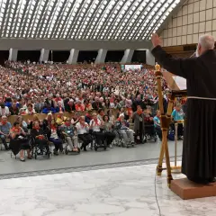 Ein charismatischer Gemeinde-Leiter mit erhobenen Armen spricht zu einer riesigen Menge von Gläubigen, die in einem großen Auditorium sitzen.