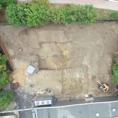 Wykop archeologiczny na placu budowy, gdzie prowadzone są badania przed pozwoleniem na budowę. Widoczny koparka i ślady dawnej zabudowy.
