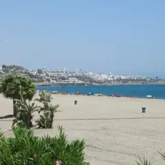 La **playa de la Mena** se extiende con arena dorada y palmeras. Al fondo, un pueblo blanco se asoma al mar azul.