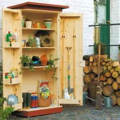 Ein selbstgebauter Gartenschrank aus Holz, gefüllt mit Gartenutensilien wie Töpfen, Werkzeugen und Gummistiefeln. Daneben liegt aufgeschichtetes Brennholz.