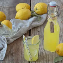 Frische Zitronen, ein Glas mit Eis und Strohhalmen, sowie eine Flasche selbstgemachter Zitronensirup auf Holzhintergrund.