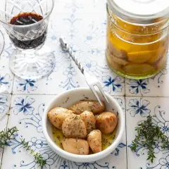 Ail confit au four, tendre et parfumé, servi dans un bol avec des herbes. Deux verres de vin rouge et des bocaux d'ail confit complètent la scène.