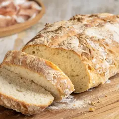 Pane fatto in casa, croccante fuori e soffice dentro, tagliato su un tagliere di legno. Sullo sfondo, un piatto di affettati.
