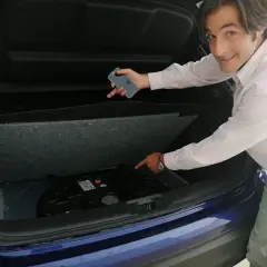 Hombre señala el espacio vacío en el maletero de un coche azul, donde debería estar la rueda de repuesto. "He pinchado y no tengo rueda de repuesto", parece decir.