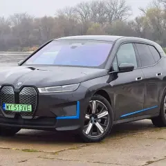 Ciemnoszary BMW iX z niebieskimi akcentami stoi nad rzeką, z mostem w tle.