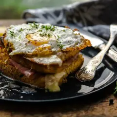 Ein üppiger Croque Monsieur mit Spiegelei, Schinken und geschmolzenem Käse, garniert mit Kräutern und Beilagen.