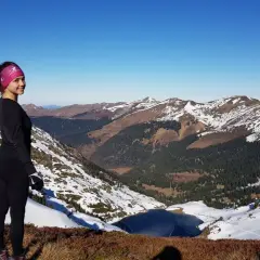 Vue panoramique depuis le pic du lion. Une femme souriante, en tenue de sport, admire le paysage montagneux enneigé et un lac gelé.