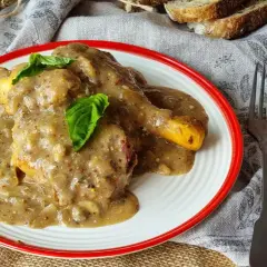 Delicioso pollo a la mostaza cubierto con una cremosa salsa, servido en un plato blanco con borde rojo. Acompañado de pan y cubiertos.