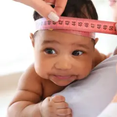 Ein Baby mit dunkler Haut wird der Kopfumfang gemessen. Ein Maßband zeigt etwa 40 cm.