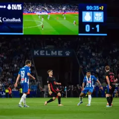 Piłkarze na boisku, jeden z nich w czarnej koszulce z numerem 19, próbuje odebrać piłkę. To przykład taktyki "co to pressing" w akcji. Wynik 0-1 na tablicy.