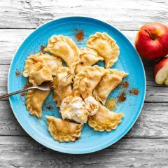 Pyszne pierogi z jabłkami, posypane cynamonem, na niebieskim talerzu, obok jabłka.