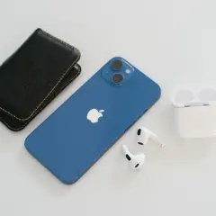 Niebieski iPhone 13, który wyszedł we wrześniu 2021, leży obok czarnego etui i białych słuchawek AirPods.
