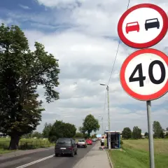 Znaki drogowe: zakaz wyprzedzania i ograniczenie prędkości do 40 km/h. Samochody jadą drogą, po prawej stronie przystanek autobusowy.