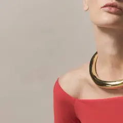 A woman wears a bold gold necklace with a curved neckline, complementing her red off-the-shoulder top.