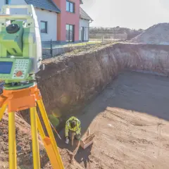 Geodeta dokonuje pomiaru rzeźby terenu na budowie. Widać niwelator i pracownika w wykopie.