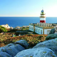 Faro en las islas Galicia, con el mar azul de fondo y rocas cubiertas de vegetación.