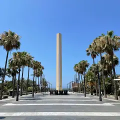 Un obelisco se alza en la plaza de las velas Almería, flanqueado por palmeras y un cielo azul despejado.