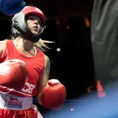 A determined female boxer in red gear and headguard prepares for a white collar boxing match, her red gloves raised.