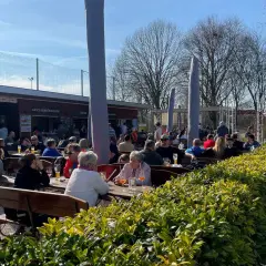 Menschen genießen das sonnige Wetter im Carles Biergarten. Gäste sitzen an Holztischen, trinken und unterhalten sich.