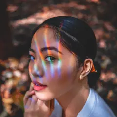 Un occhio a mandorla cattura la luce arcobaleno, riflessi colorati danzano sul suo viso mentre guarda pensierosa.