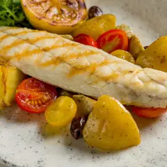 Filetto di persico grigliato su un piatto con patate arrosto, pomodorini, olive e limone caramellato.