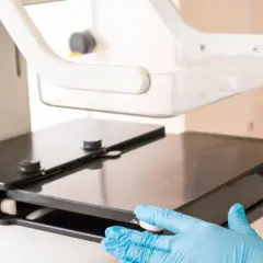 Ręce w niebieskich rękawiczkach przygotowują aparat do mammografii. Od ilu lat warto badać piersi?