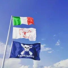 Bandiera azzurra con croce bianca, simbolo di salvataggio, sventola accanto alla bandiera italiana sotto un cielo azzurro.