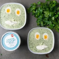 Två skålar med spenatsoppa som ser ut som ansikten, med ägg som ögon och gräddfil som mun. Bredvid står en burk grekisk yoghurt och en grön kvist persilja. Ett roligt spenatsoppa recept!