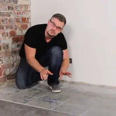 Uomo che mostra la posa pavimento pvc su pavimento esistente, con mattoni a vista e termosifone.