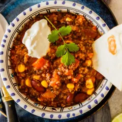 Herzhaftes Chili sin Carne mit Quinoa, Bohnen und Mais, garniert mit Sauerrahm und Koriander. Dazu eine Tortilla-Ecke und Limettenspalten.